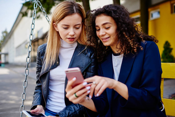 Young multiethnic ladies in casual clothes sitting on bench together and messaging on mobile in street with building on background in day - Foto, Imagen