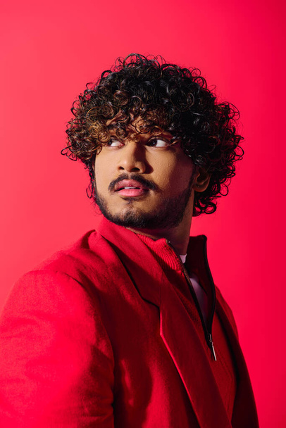 Jonge Indiase man met krullend haar showcasing rode jas op levendige achtergrond. - Foto, afbeelding