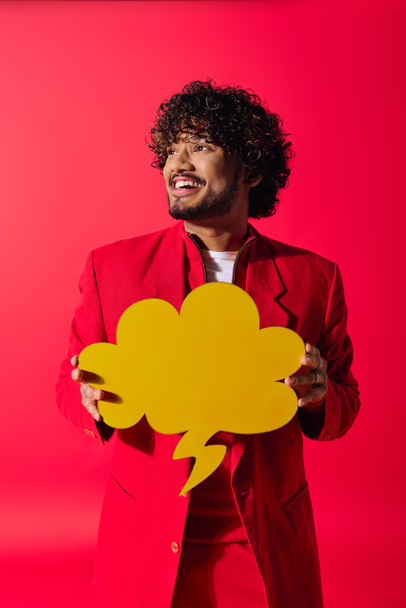 A stylish young Indian man in a red suit holding a bright yellow speech bubble. - Photo, Image