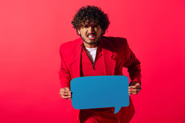 Živý mladý muž v červeném obleku drží modrou řečovou bublinu na živém pozadí. - Fotografie, Obrázek