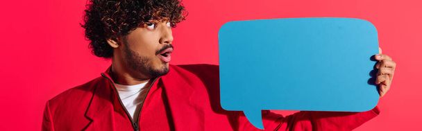 Young Indian man in vibrant red jacket holding a blue speech bubble. - Photo, Image