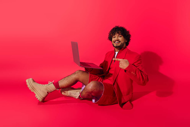 Junger indischer Mann in pulsierendem Outfit sitzt auf dem Boden und ist in Laptop-Arbeit vertieft. - Foto, Bild