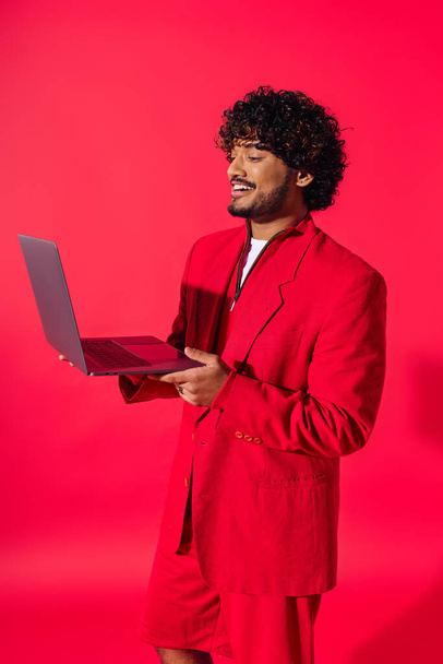 Schöner junger indischer Mann in leuchtend rotem Anzug hält einen Laptop in der Hand. - Foto, Bild
