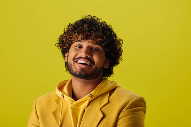 Schöner junger indischer Mann mit lockigem Haar posiert in einem gelben Kapuzenpulli vor einem lebhaften Hintergrund. - Foto, Bild