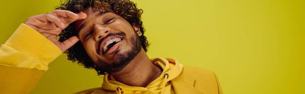 Un guapo joven indio con una vibrante sudadera con capucha amarilla hace una expresión divertida contra un telón de fondo llamativo. - Foto, imagen