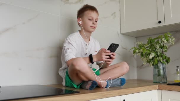 Młody chłopiec siedzi wygodnie na kuchennym blatze, pochłonięty słuchaniem muzyki przez białe słuchawki, podłączony do swojego przenośnego smartfona. Naturalne światło wypełnia pokój, podkreślając casual - Materiał filmowy, wideo