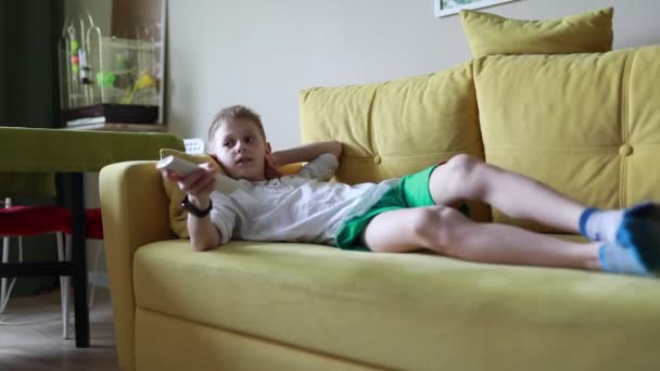 Un jeune garçon est montré allongé sur un canapé tout en tenant une télécommande dans sa main. Il semble concentré sur l'appareil à sa portée, affichant un moment de concentration et de détente. - Séquence, vidéo
