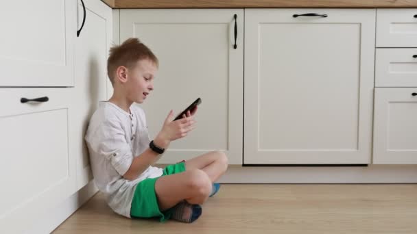 Een jonge jongen zit op de vloer, verzonken in zijn mobiele telefoon scherm. Zijn vingers tikken en vegen als hij interageert met het apparaat, volledig ondergedompeld in de digitale wereld. - Video
