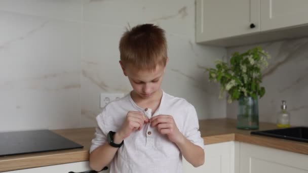 Młody chłopiec ze skupieniem uwagi uczy się zapiąć koszulę w zaciszu słonecznej, nowoczesnej kuchni. Jego małe palce umiejętnie pracują, aby zabezpieczyć każdą dziurkę od guzika, zaznaczając kamień milowy w rozwoju - Materiał filmowy, wideo