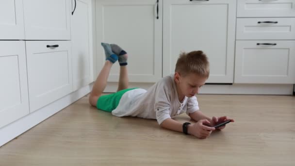 A young boy lies comfortably on his stomach on a bright, airy kitchen floor, engrossed in playing games or browsing through a smartphone. He is dressed in casual home attire, suggesting a leisurely - Footage, Video