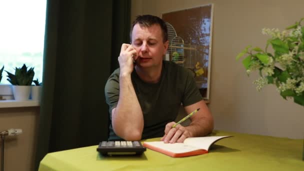 Um homem adulto está sentado em uma mesa coberta com documentos financeiros, uma calculadora e um bloco de notas. Ele aparece envolvido em uma conversa séria por telefone, possivelmente com um provedor de serviços ou - Filmagem, Vídeo