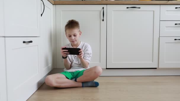 Een gefocuste jongen zit op de keukenvloer met zijn benen gekruist, diep verdiept in het spelen van een spel op zijn handheld console, met zonlicht binnenstromend uit de kamer achter hem. - Video