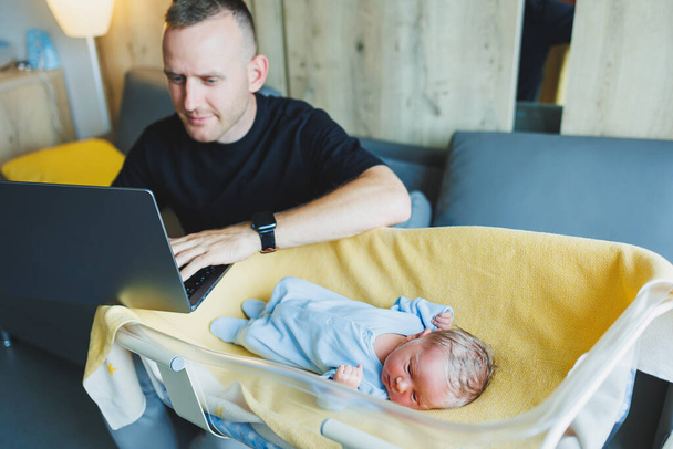 Un jeune père travaille sur un ordinateur portable et s'assoit à côté d'un bébé. Travailler à la maison avec un petit enfant - Photo, image