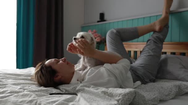 Als het zonlicht de kamer zachtjes verlicht, deelt een vrolijke huisdier eigenaar liefdevolle speeltijd met hun kleine witte hond op het comfort van een gezellig bed, wat wijst op een nauwe band tussen hen. - Video