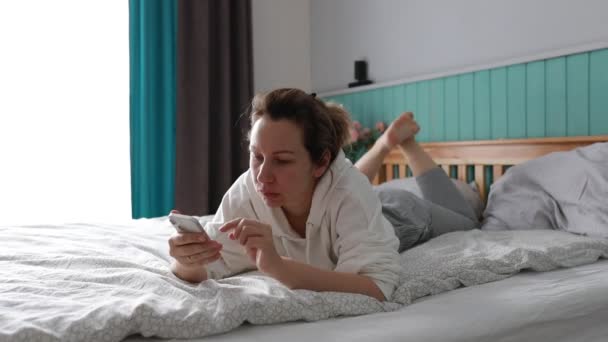 Een jonge vrouw geniet van een ontspannen ochtend, liggend in haar comfortabele bed als ze terloops bladert door haar smartphone, mogelijk inhalen op berichten of social media-updates in het zachte daglicht - Video