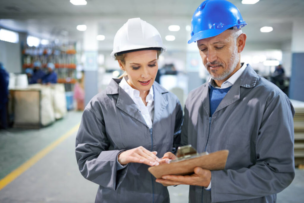 Man, woman and conversation with clipboard in factory for quality inspection or safety checks and process monitoring. Team, communication and board for manufacturing information in printing warehouse. - Photo, Image
