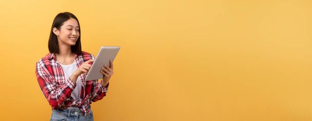 Una giovane donna asiatica allegra in camicia a quadri sta usando un tablet digitale su uno sfondo giallo vibrante con spazio per la copia - Foto, immagini