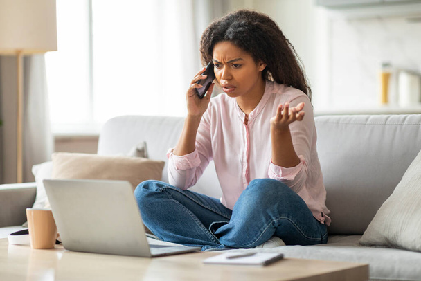Czarna kobieta wydaje się zaniepokojona podczas rozmowy przez telefon i patrząc na ekran laptopa, podkreślając poważną rozmowę w domu - Zdjęcie, obraz