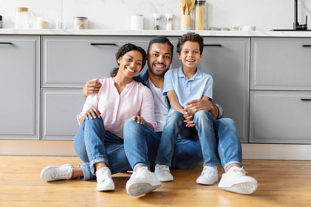 Чорні батьки і дитина сміються разом, сидячи на підлозі на кухні, демонструючи спокійну сімейну атмосферу - Фото, зображення