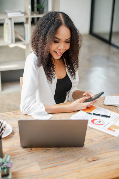 Glimlachende jonge vrouw in een witte blazer zit aan een houten bureau, werkt aan een laptop en houdt een smartphone vast. - Foto, afbeelding