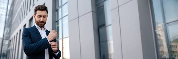 Улыбающийся уверенно спокойный привлекательный молодой кавказский мужчина гендиректор с бородой в костюме возле современного офисного здания, на открытом воздухе, копировальное пространство. Успешный бизнес, бизнесмен в городе, реклама, босс и карьера - Фото, изображение