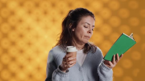 Tired woman reading book, holding cup of coffee, isolated over studio background. Exhausted bookworm enjoying novel and caffeine beverage, lacking energy, struggling to stay awake, camera A - Footage, Video