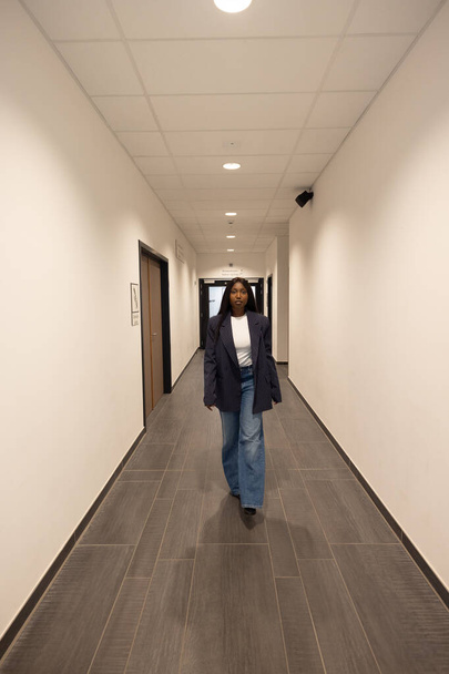 中型の建物を持つアフリカ系アメリカ人女性は,明るいオフィスの廊下でカメラに向かって歩くのが特徴です. 彼女はリラックスした姿勢を持ち,自信を伝えます. 彼女のスタイルはカジュアルなプロフェッショナル - 写真・画像