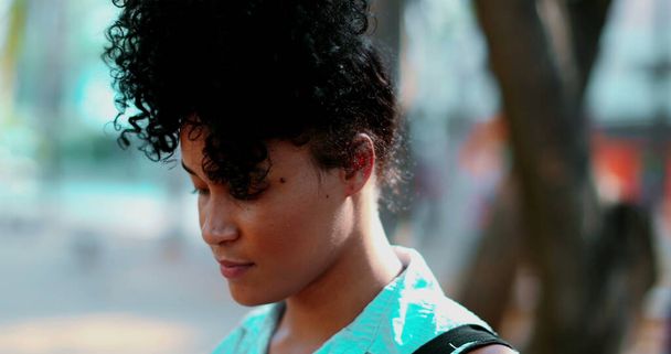 Πορτρέτο ενός Pensive νεαρή μαύρη γυναίκα στέκεται υπαίθρια σε ειρηνική διαλογιστική κατάσταση, close-up πρόσωπο στην παρακολούθηση πυροβόλησε κατά τη διάρκεια της ηλιόλουστης ημέρας και στοχαστικό συναίσθημα - Φωτογραφία, εικόνα