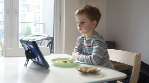 Młody chłopiec siedzi przy stole jadalnym, uważnie obserwując tabliczkę podpartą przed nim, jedząc śniadanie w słonecznej kuchni. Dziecko sporadycznie wchodzi w interakcje z tabletką - Materiał filmowy, wideo