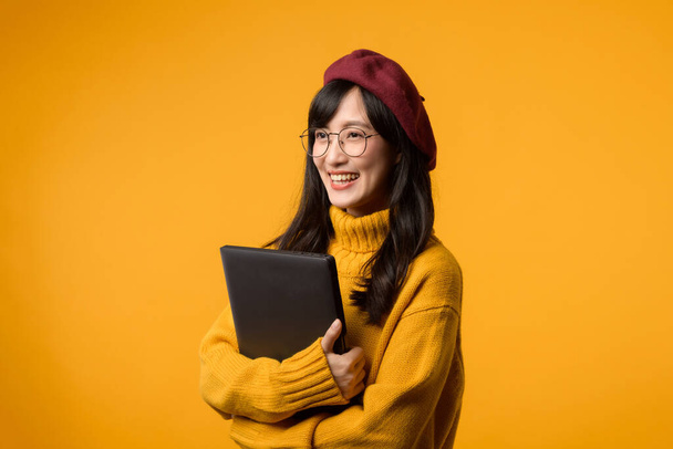 Sullo sfondo giallo sole, una giovane donna asiatica utilizza con fiducia il suo computer portatile mentre indossa un maglione giallo e un berretto rosso. - Foto, immagini