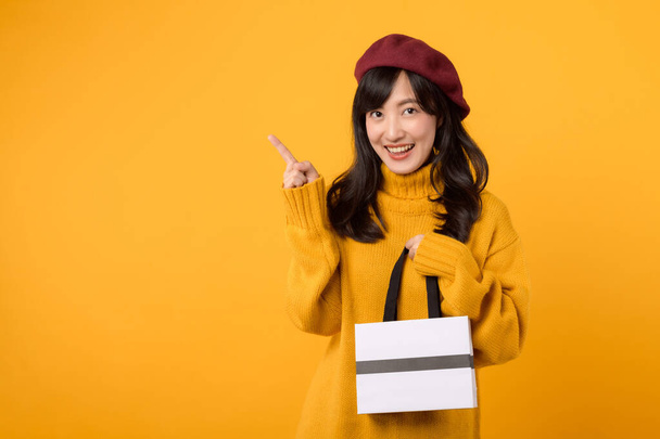Junge Asiatin um die 30, in einem gelben Pullover und roter Baskenmütze vor gelbem Hintergrund, hält eine Einkaufstasche und zeigt mit dem Finger. - Foto, Bild