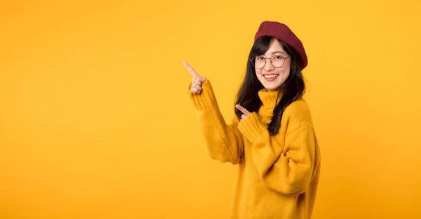 Stylische Frau in ihren Dreißigern, trägt einen gelben Pullover, rote Baskenmütze und Brille und zeigt mit dem Finger auf freien Kopierraum vor leuchtend gelbem Hintergrund.. - Foto, Bild