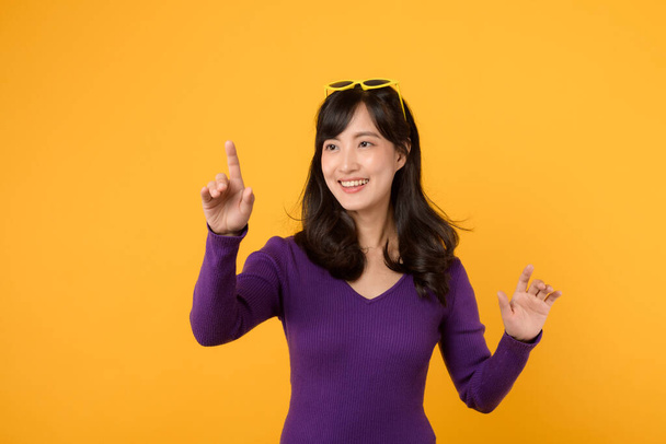 黄色の背景に隔離された仮想スクリーンに触れる頭の上に紫色のシャツと黄色のサングラスを着用する若いアジアの女性. 技術コンセプト. - 写真・画像