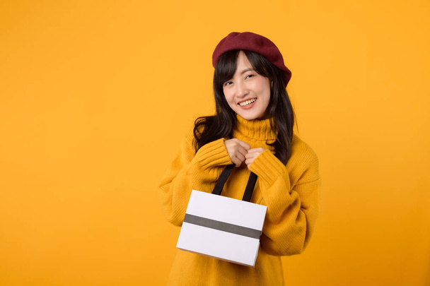 Jeune femme asiatique trentaine, avec un sac en papier à la main, portant un pull jaune et un béret rouge, célébrant son achat sur un fond jaune. - Photo, image