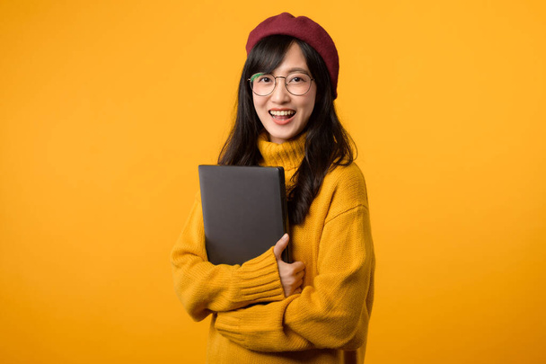 Egy fiatal ázsiai nő laptoppal, sárga pulóverben és piros svájcisapkában, kiemelkedik élénk sárga háttérrel. - Fotó, kép