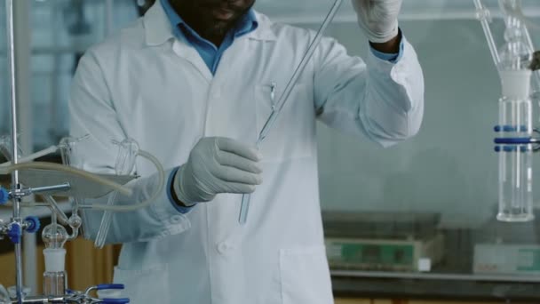 Cropped midsection shot of African American male laboratory technician in white coat and gloves filling tube with light blue liquid from glass straw, stirring in circular motion, putting down on rack - Footage, Video
