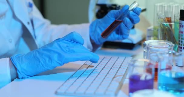 Científico en guantes azules trabaja en la computadora sosteniendo tubo de ensayo con líquido rojo. Análisis de sangre o líquidos tóxicos - Metraje, vídeo