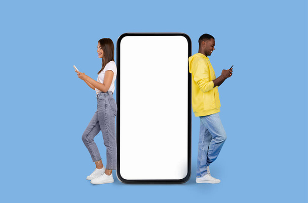 A young woman and a black man stand back-to-back, engrossed in their smartphones, beside a large blank smartphone screen on a blue background - Photo, Image