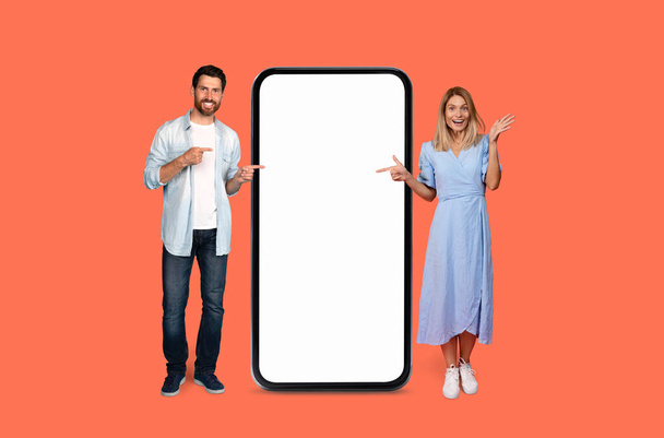 Muž a žena stojí po obou stranách přerostlého smartphonu s prázdnou obrazovkou, ukazují a vypadají vesele - Fotografie, Obrázek
