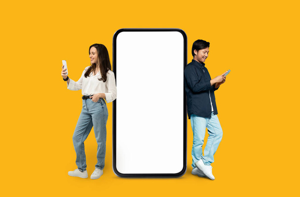 Dois amigos Multiracial homem e mulher interagindo com um grande smartphone com tela vazia em um fundo laranja - Foto, Imagem