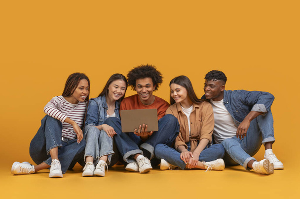 Połączony zespół młodych, wielonarodowych przyjaciół zajmujących się laptopem, symbolizujący technologię w kontekście przyjaźni, odizolowany na żółtym tle - Zdjęcie, obraz