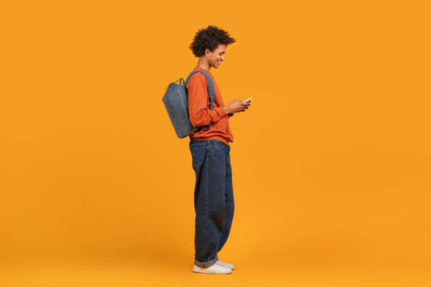 Un hombre negro moderno en ropa casual con mochila usando un teléfono inteligente, enviando mensajes de texto a amigos, aislado sobre fondo naranja - Foto, Imagen