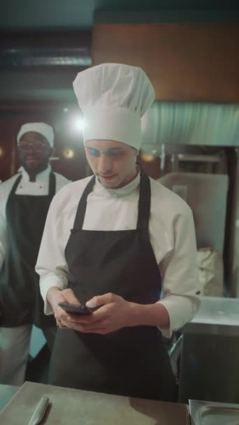 Pionowe ujęcie kucharza w fartuchu i kapeluszu stojącego w kuchni restauracyjnej, korzystającego z telefonu, witającego kolegę z zapięciem ręcznym i rozmawiającego z nim - Materiał filmowy, wideo