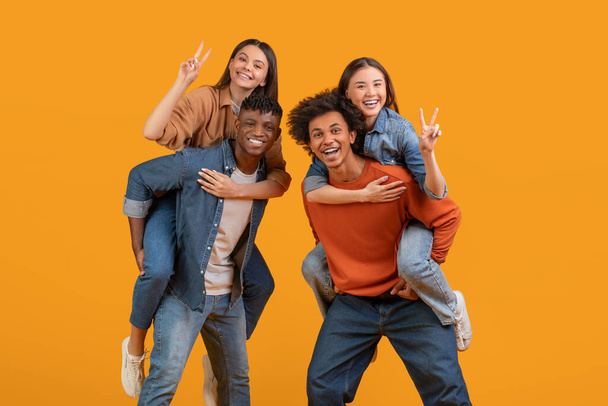 A joyful multiethnic group of friends piggybacking, showing peace signs, and laughing, isolated on an orange background portraying happiness and team spirit - Photo, Image