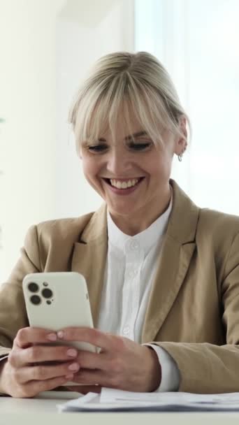 Happy Caucasian female office worker using phone and laughing spending work break in the office. Relaxing and browsing internet. Vertical video. - Footage, Video
