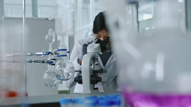 Střední záběr mladé čínské vědkyně v bílém laboratorním plášti a rukavicích se ohýbá, hledí do mikroskopu, upravuje zvětšení v okuláru a studuje vzorek - Záběry, video