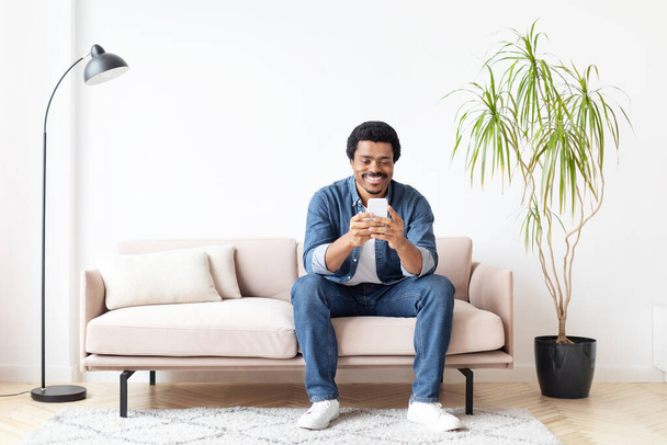 Радостный африканский американец дома пишет смс на своем смартфоне, выражая общение и счастье - Фото, изображение