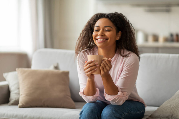Giovane donna nera positiva seduta da sola sul divano a casa, che beve caffè a occhio chiuso e sorride. Stile di vita Millennials - Foto, immagini