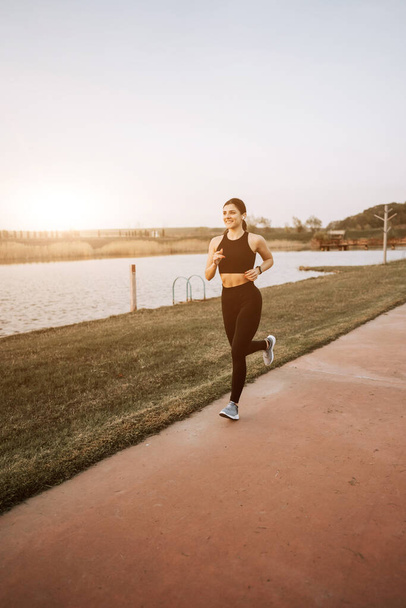 Nuori nainen urheiluvaatteissa juoksee pitkin pengertä auringonlaskun aikaan. Terveellisen elämäntavan käsite - Valokuva, kuva