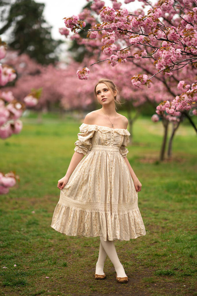 Bella giovane ragazza bionda in abito vintage in pizzo in piedi nel parco primaverile vicino fiori di fiori rosa. Tenderness modello romantico in posa. - Foto, immagini